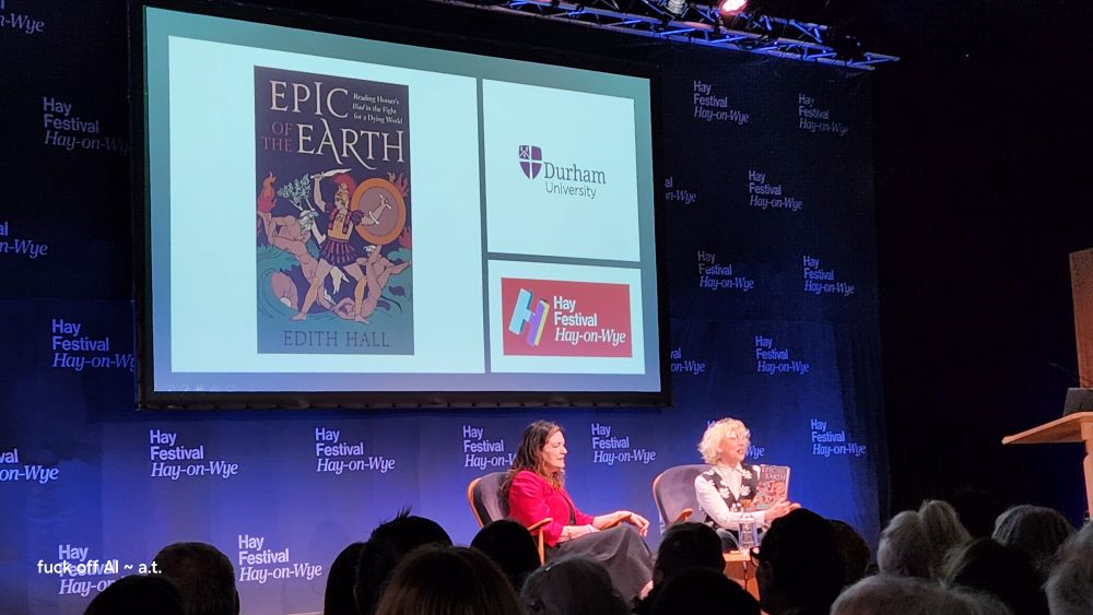Photo of panel stage with two women in chairs chatting with each other. Behind them is a large screen showing the illustrated cover of a book titled EPIC OF THE EARTH by Edith Hall. 