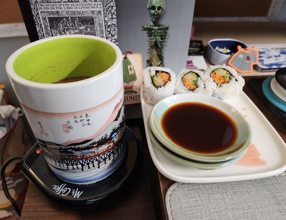 Photo of a ceramic square plate with soy sauce and sushi on it. There is also a white mug with an illustrated bridge on the outside and green coloring on the inside. In the background are postcards set up as decor of an ancient skeleton on display and next to that a postcard with an illustration from an old book of arthurian lore. Three men ride horses, heading to adventures unknown. Text above them: The story of the glittering plain or the land of living men. Upper right of image there is a tiny decal of the character K9 from the show 