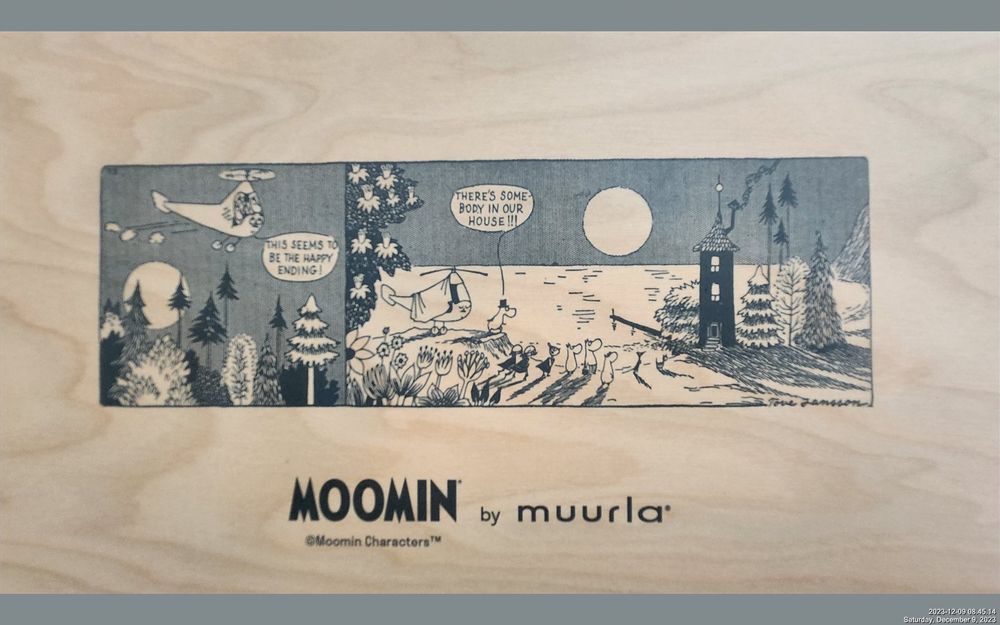 One side of a wooden tray with a black and while graphic printed on it showing two illustrated panels featuring a family called The Moomins. In the first panel they are flying in a chopper over a bucolic little forest. In the next panel they are standing on a ridge looking at a little round tall house with smoke coming out of the chimney. It is set by a shoreline and a little pier extends into the water. A full sun is near the horizon, casting shadows behind all the characters. Dialogue from the main character, Moomin, who looks kind of like a humanized hippo in first panel in the helicopter: This seems to be the happy ending! Second panel: There's somebody in our house!