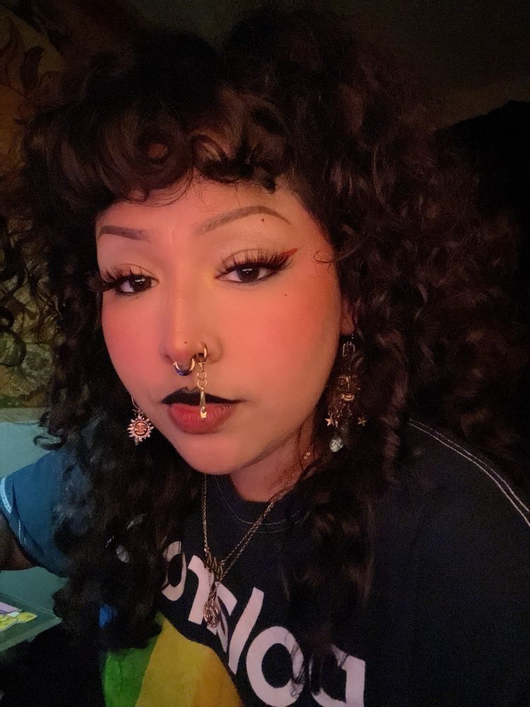 selfie in semi golden lighting. i have on all gold jewelry such as sun and moon dangly earrings, septum and a dangle hand nostril + a 444 pendant necklace coupled with a sun pendant necklace. my shirt is a rainbow polaroid tee. my hair is up in a hair pony which has my curly brown hair frame my face nicely. my eyeliner is red + thick lashes and gold inner corner. 