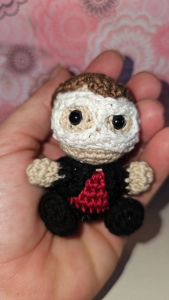 Amigurumi de Chris, un personaje de Mi espíritu y tu voz. Cabe en la palma de una mano, y va vestido de negro, con camisa roja y una máscara que le cubre el rostro