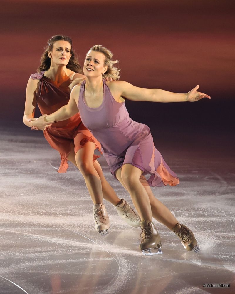 Gabriella Papadakis and Madison Hubbell performing their number at Art on ice 2025
Gabriella is wearing an orange dress that fades into purple near the strap, Madison is wearing a pastel purple dress with a little bit of orange in the back of her dress