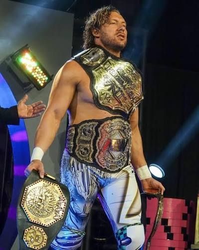 Kenny Omega wearing the TNA World Heavyweight Championship belt around his neck and left shoulder, the AEW Men's World Championship belt around his waist, carrying the IMPACT World Championship belt in his left hand and the AAA Mega Championship belt in his right hand.