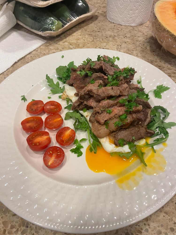 A plate of sliced steak on top of a couple of fried eggs on a bed of arugula, sprinkled with torn chervil leaves, with halved cherry tomatoes off to the side as a garnish. Some yolk is leaking out onto the plate from underneath one of the eggs.