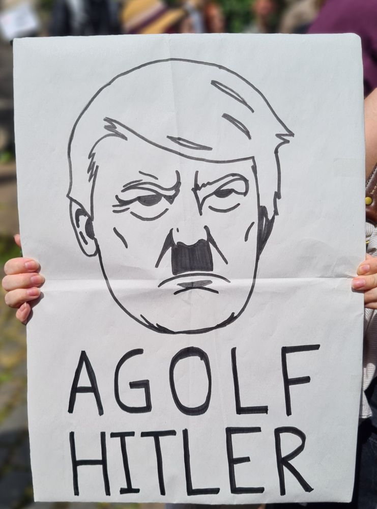 White paper sign held up in front of blurred background of streets paved with setts and indistinct crowd, black line art is Donald Trump caricature with Hitler moustache saying AGolf Hitler at bottom of sign