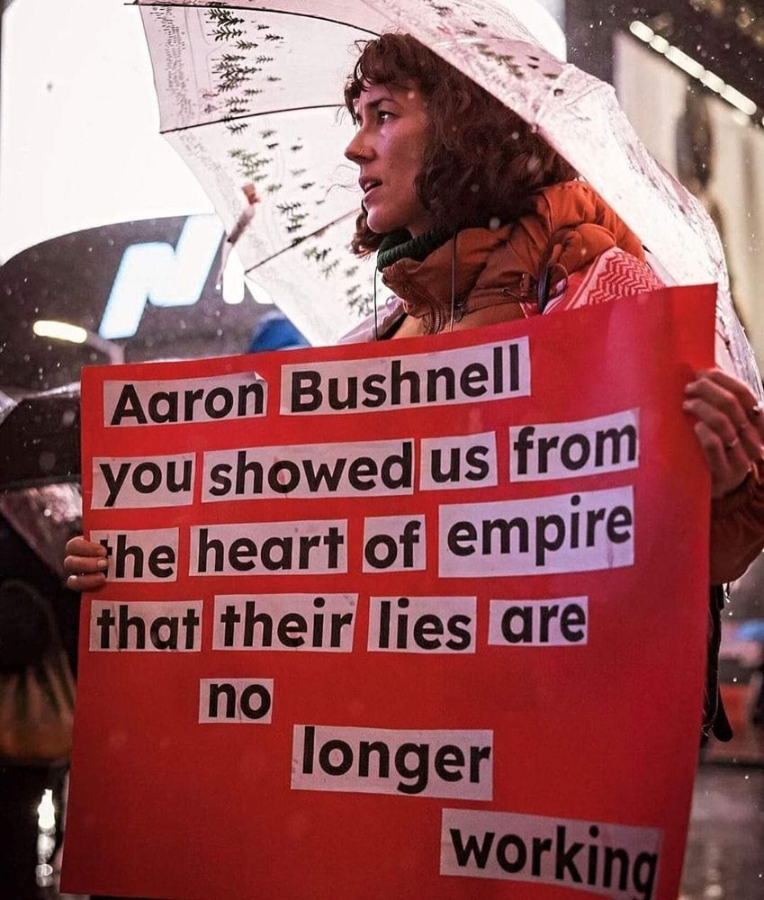 Person holding sign stating "Aaron Bushnell you showed us from the heart of empire that their lies are no longer working"