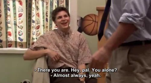 George Michael sits on a chair in a small, brightly lit room, smiling nervously up at someone offscreen. He wears a buttoned-up, short-sleeved shirt with a muted print. Behind him, there’s a window with cartoon-printed curtains and a wall-mounted basketball hoop. Michael stands in front of him, seen from the waist down, wearing a blue shirt with rolled-up sleeves and khakis. The mood is awkward, with George Michael’s response highlighting his lonely nature.