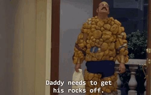 Tobias stands proudly in the hallway wearing a bulky, ridiculous rock-covered bodysuit resembling The Thing from *Fantastic Four*. His mustache and bald head are visible above the costume, and he holds a plastic bag in one hand. He declares with misplaced confidence, "Daddy needs to get his rocks off," completely unaware of the double entendre.
