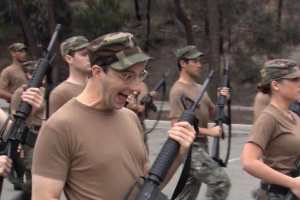 Buster marches in a military drill formation, gripping a rifle with exaggerated enthusiasm. His mouth is wide open in a giddy, almost manic smile, and his eyes are wide behind wire-rimmed glasses. He wears a camo cap and brown T-shirt like the others, but his over-the-top expression starkly contrasts with the serious, focused demeanor of the soldiers around him.

