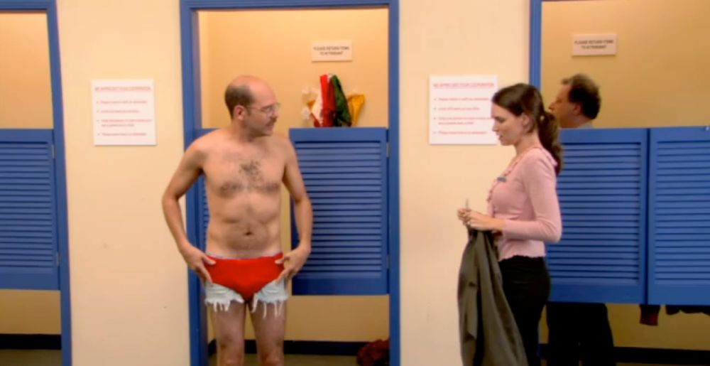 Tobias stands shirtless in a changing room, awkwardly showing off a pair of cut-off jean shorts with bright red briefs pulled high over them. He looks toward a disinterested saleswoman holding a blazer, her expression blank as she avoids eye contact. In the background, another man peeks into a fitting room mirror. Tobias’s stance is proud despite his ridiculous outfit.