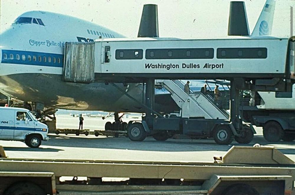 A Pan Am Boeing 747, named *Clipper Belle of the Sky*, is parked on the tarmac at Washington Dulles Airport. A unique mobile lounge, marked with the airport’s name, is docked to the aircraft for boarding or deplaning. The mobile lounge is elevated, with stairs and support structures beneath it. Ground crew and vehicles, including a white van and service carts, are active around the plane, suggesting a busy operational moment.

