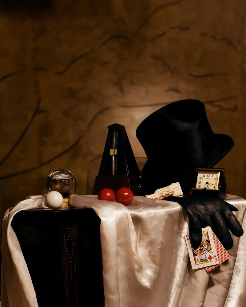 Photograph taken by the brilliant photographer Kenneth Lam. It is taken in “The Roses of Elagabalus” - a new queer clubhouse in Dalston. In the background a beautiful minimalist mural of a reclining nude figure. In the foreground a still-life of props from Neil Kelso’s show. From left to right a white billiard ball, a crystal ball, a metronome, two red billiard balls, some dollar bills, a top hat, a leather glove gripping two playing cards, and a vintage clock. It is arranged on a cream coloured satin cloth, on a vintage wooden table with a diamond pattern on its leg. The lighting is so perfect that it looks like a beautiful renaissance painting. It has a magical and mysterious atmosphere.