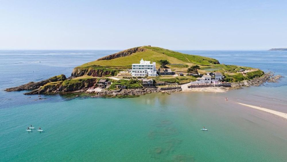 Photograph of the beautiful Art Deco Burgh Island Hotel and the tiny charming ancient Pilchard Inn, on Burgh Island. The tide has cut the island off from the mainland (Devon, UK) and the sun is shining.