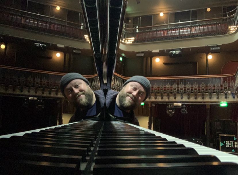 Neil Kelso a smiley white man with a brown beard. He is wearing a grey woolen hat and blue jacket. He is peering out from behind a grand piano. The piano keys and his face are reflected in the shiny black polished piano. In the background is the balcony and dress circle of Hoxton Hall.