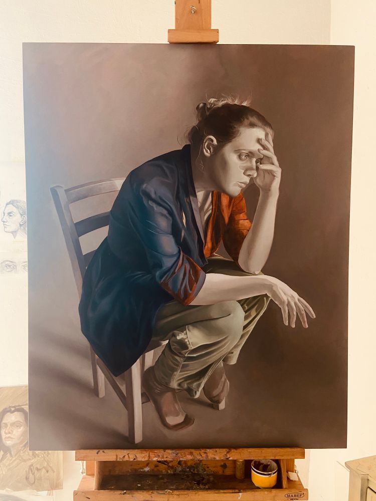 An unfinished painting sits on an easel. The figure in the painting is a woman seated on a chair with her hand to her head. She is wearing a black top and light green pants, but the rest of the painting is in gray scale. 