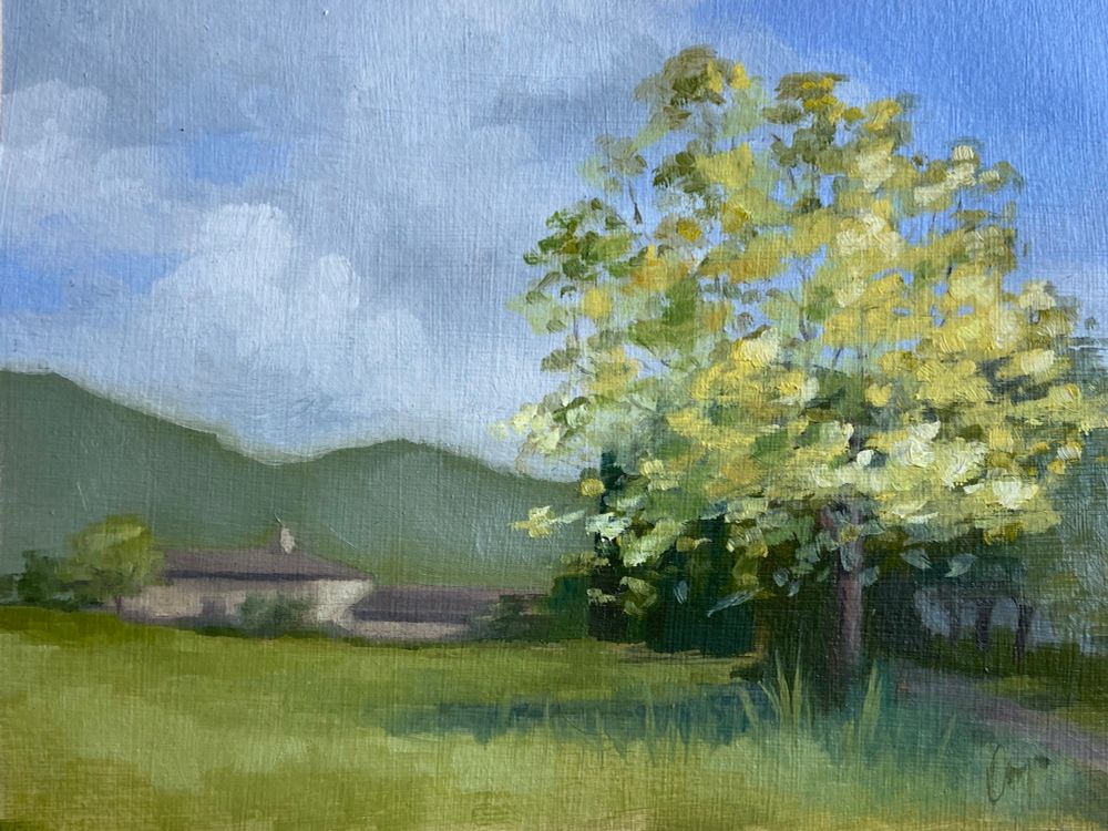 An oil painting of a landscape. In the background is a beige building in front of large mountains and a blue sky. In the foreground is a large grassy field. To the right a path is barely visible, it is lined with trees. Between the oath and the field is a large tree with yellow and green leaves.
