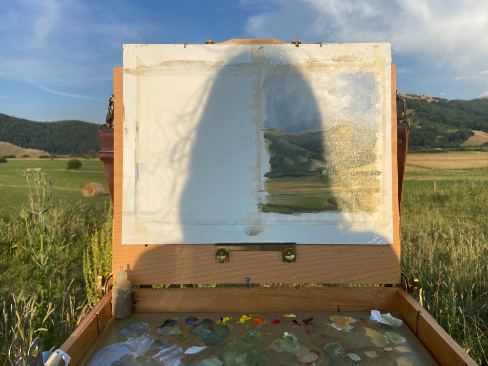 The shadow of an artist falls on an easel with a landscape painting on it. At the bottom of the image, a portion of the artists palette is visible. Behind the easel is a large open field with mountains in the background. 