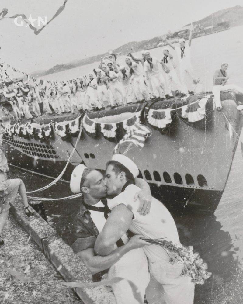 a staged black and white photo of old-timey dressed sailors. In the foreground, two male sailors are passionately kissing, one holding flowers. In the background stands dozens of sailors cheering and waving.

Photo posted by Gay Star News. Details on the artwork: A staged recreation of sailors celebrating Victory Day in 1945, including two men kissing in the right foreground. Photo was taken by David LaChapelle for Diesel Jeans in 1994. This photo is currently on display at The Getty Museum in the exhibit “Queer Lens” at the Getty Centre, Jun 17–Sep 28, 2025