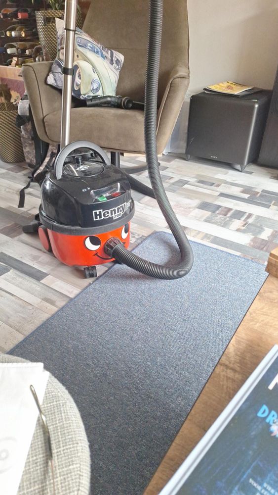 Stofzuiger (Henry-stofzuiger) met een vrolijk gezicht op het apparaat, staand op een blauwgrijs vloerkleed en houten vloer. Op de achtergrond staat een bruine fauteuil met een kussen waarop een afbeelding van een gele auto is te zien. Daarnaast zie je een wijnrek, een plant, en rechts van de stoel twee grote zwarte speakers. Op een kastje liggen tijdschriften en er staan twee gekleurde uilenbeeldjes. Het geheel oogt als een gezellig ingerichte woonkamer.