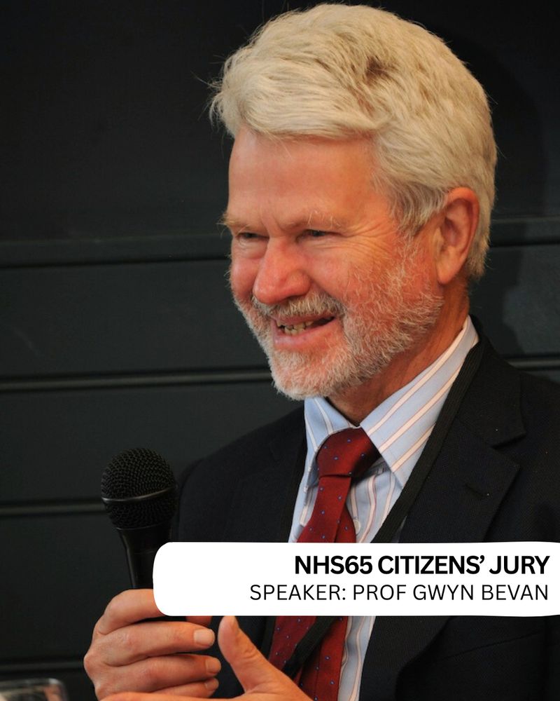 Colour photograph of Prof Gwyn Bevan, he is looking off camera, holding a microphone with both hands, he is smiling. He is wearing a black suit, white and blue striped shirt and dark red tie.