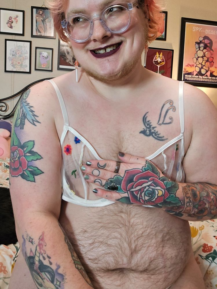 Bee wearing dark lipstick and clear glasses, with a white bra that has flowers in different colors. They're holding the bra on with one hand and smiling, looking off camera