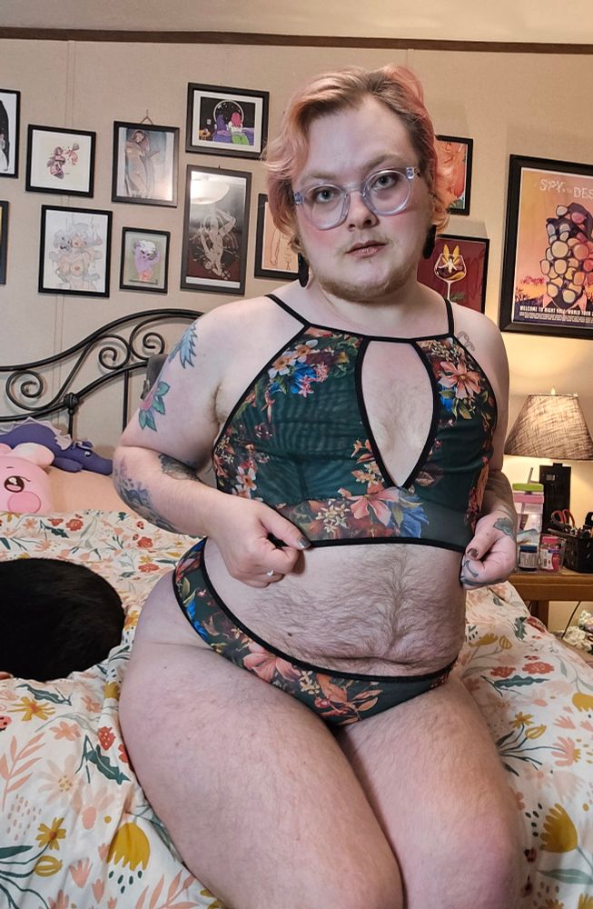 Bee sits on their bed with their knees to the side, wearing a dark green floral two piece lingerie set. You can see a black cat laying beside them. 