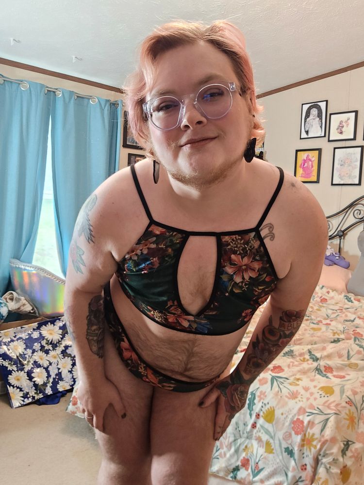 Bee leans toward the camera in a two piece lingerie set that is dark green and floral. They're wearing black cat earrings and clear frame glasses. You can see their blue curtains and floral bedspread behind them