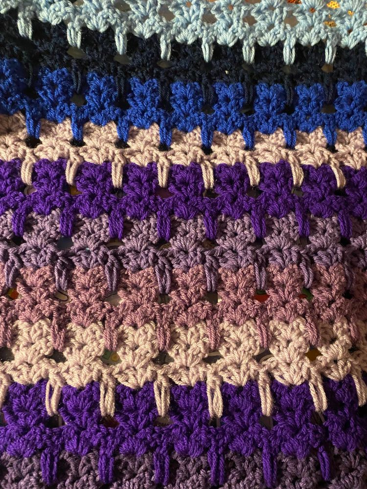Close up shot of a crochet blanket, worked in the cat stitch. Top part is made of rows in varying shades of blue, and bottom part is made of rows in varying shades of purple