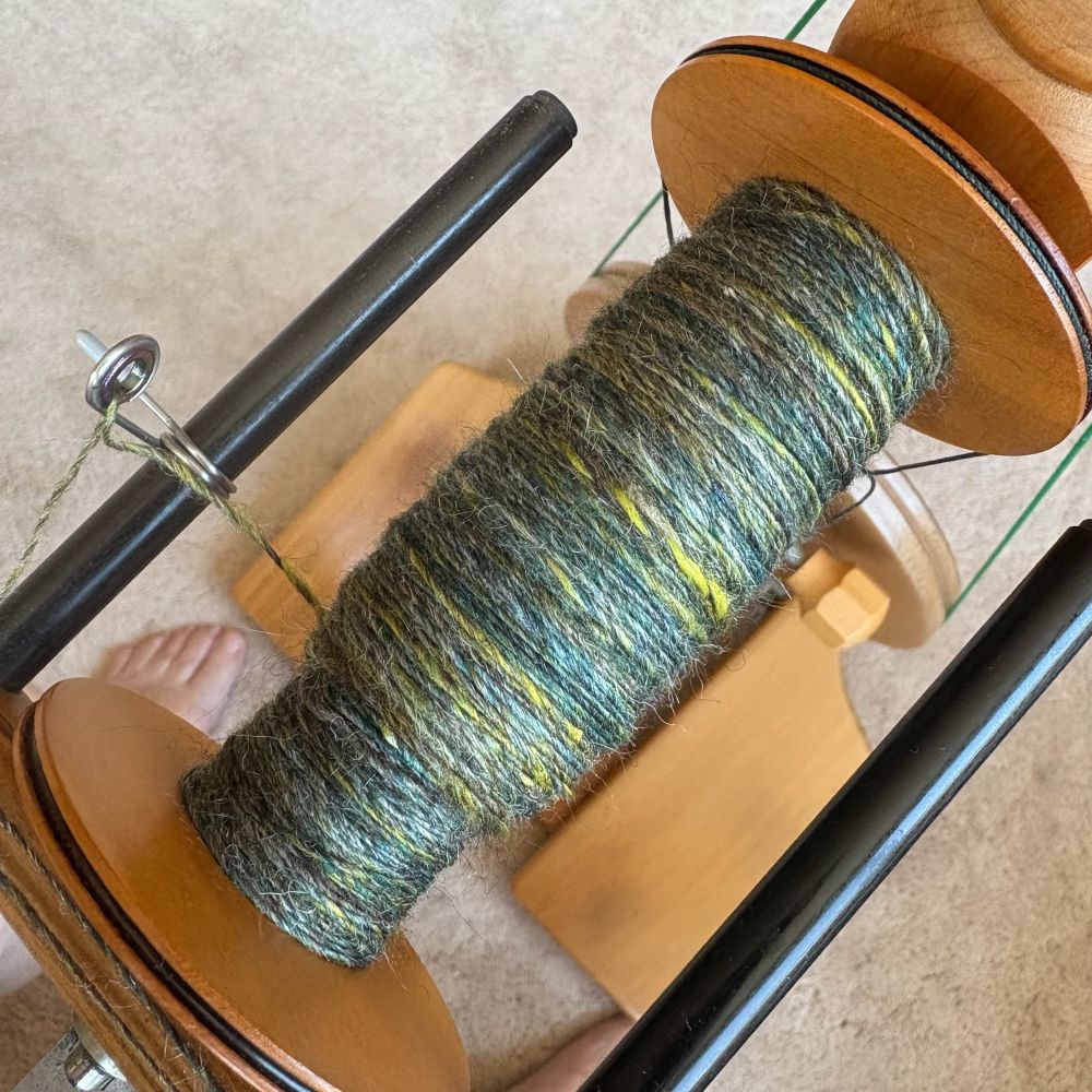 Shades of green fibre being spun, shown on a bobbin of a Little Gem spinning wheel. 