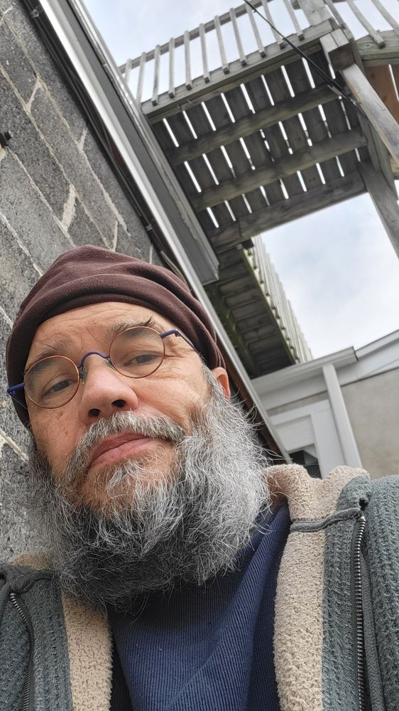 Selfie with my one dimple sitting on a staircase below other parts of the wooden staircase, next to a brick wall 