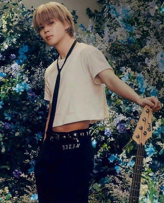 pretty jimin in a short white t-shirt that shows his sides and belly with a black loose tie around his neck, blonde hair, black pants and a black and silver belt. he is surrounded by flowers and holding the tip of an electric guitar