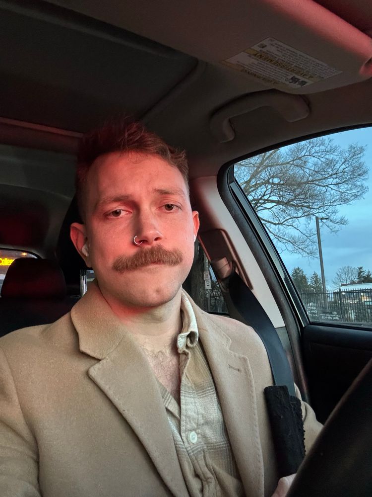 A selfie of apaulieg parked in his car. He is wearing a beige shirt and a beige jacket.