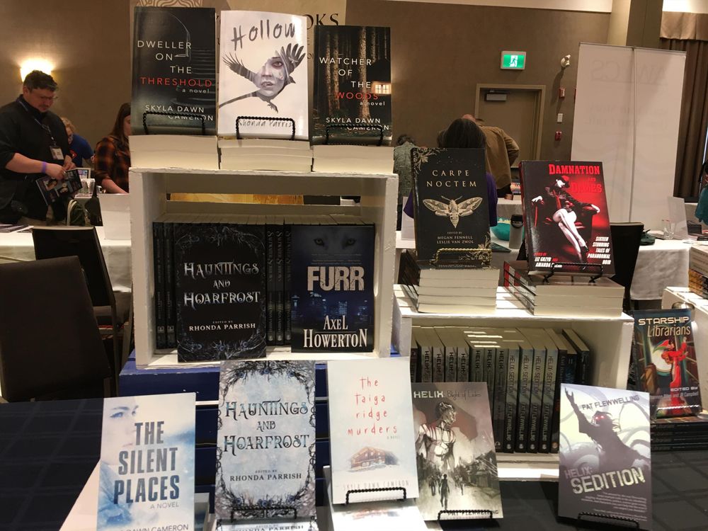 Book display at a conference. On the table are a few crates displaying books. These volumes are horror and dark fantasy. Many titles are by Skyla Dawn Cameron. There are also a few anthologies, some shapeshifter books, and YA horror.