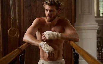 imagem da série the forsytes, um homem branco de cabelos claros e barba. ele está sem camisa, com as mãos enfaixadas.