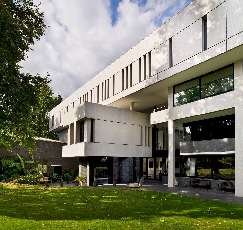 A side view of the exterior of the RCP. A long top floor clad in white tile and marked by slit windows sits above large windows and a box-like room jutting out. The grass lawn is in the foreground.