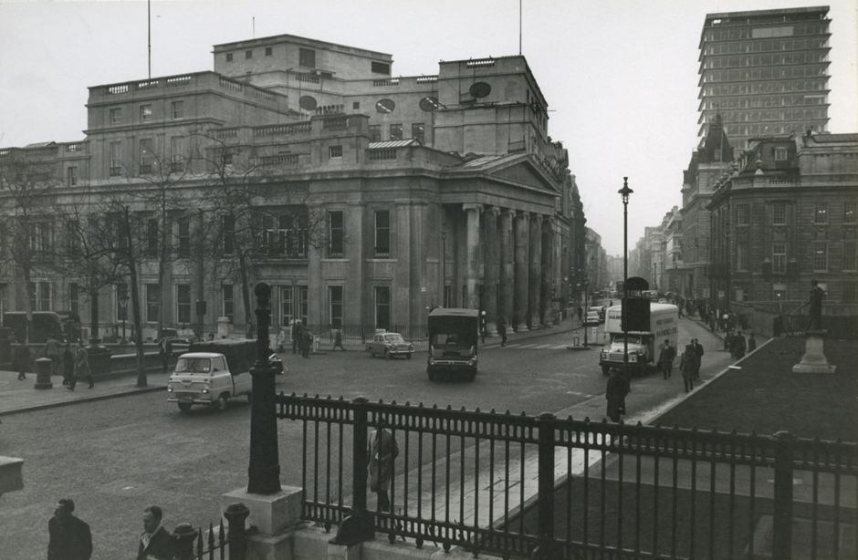 A 20th century photo of the building at Pall Mall East. You can still see the Georgian style building with sash windows and low roofs and the 6 ionic columns holding up the triangular pediment above the entrance. However larger buildings loom up behind and around and the road is populated with cars and vans and people in early 20th century dress.