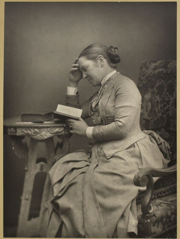 A black and white photo of Elizabeth Garrett Anderson. She is pictured in a dress, her hair in a bun, reading a book leaning her head on her hand.