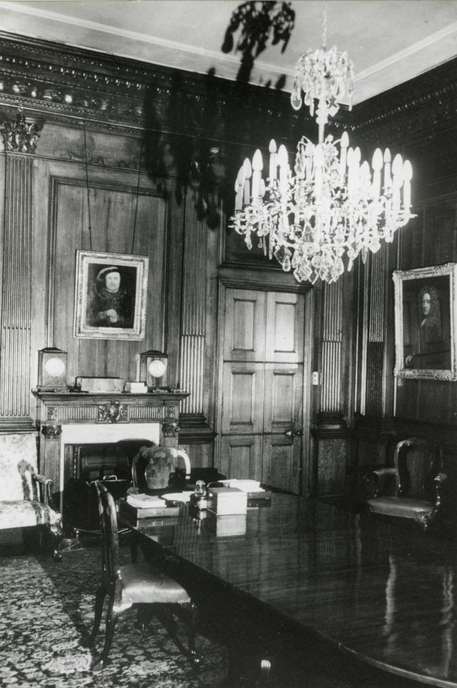 A black and white photo of the Censors room in Pall Mall East showing the Spanish Oak panelling on the walls, a central wooden desk with one chair, a chandelier above the table, a fireplace on one wall with a portrait of Henry VIII above. Other paintings and chairs can be seen around the edges of the room.