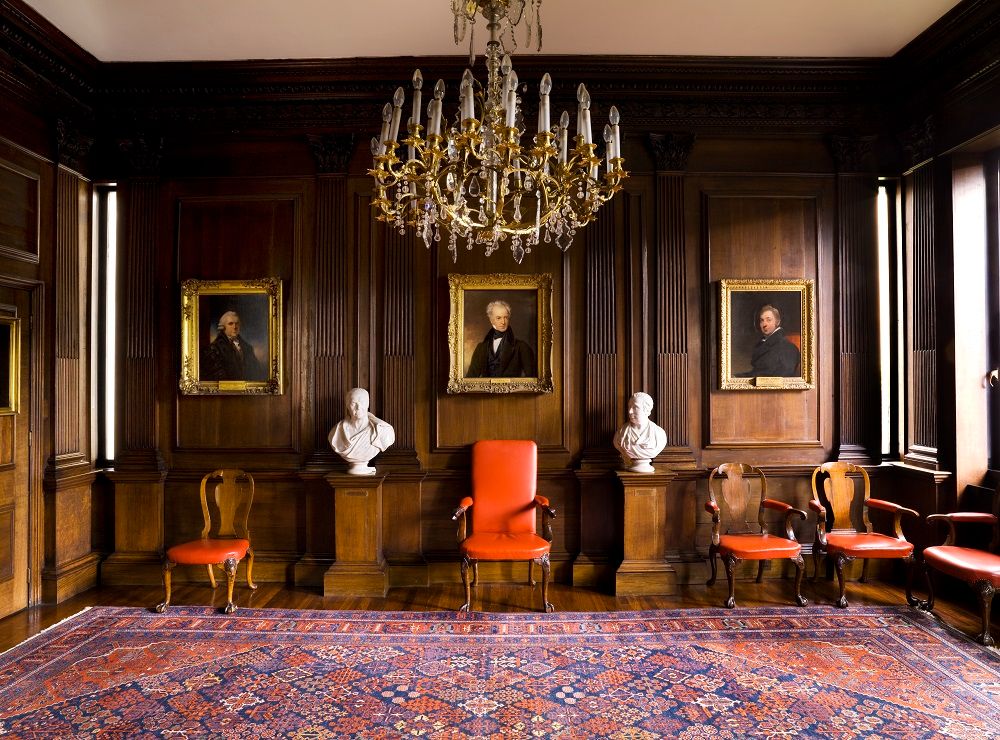 A colour photo of the Censors Room in the RCP by Regents Park today. The Spanish oak panelling liens the walls, a chandelier hangs int eh centre above and portraits, chairs and busts line the wall. 