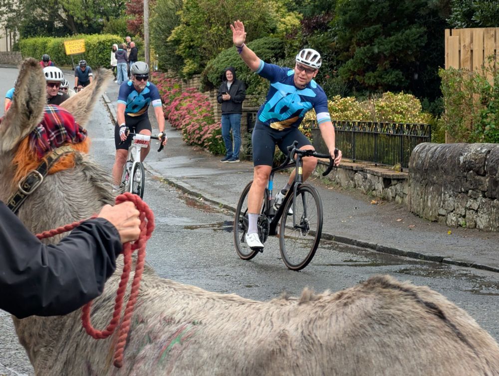 Sir Chris Hoy on a charity ride waving with a donkey in the foreground. 