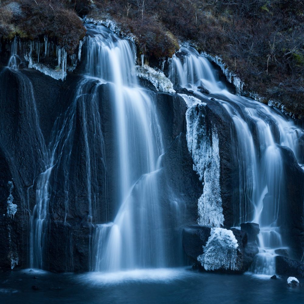 A square photograph of a waterfall, some of the surrounding rocks are rusted with ice.  The texture of the water appears soft from a long expsosure.  There is a notable blue cast to the image from It being photographed with the entire scene in shadow.