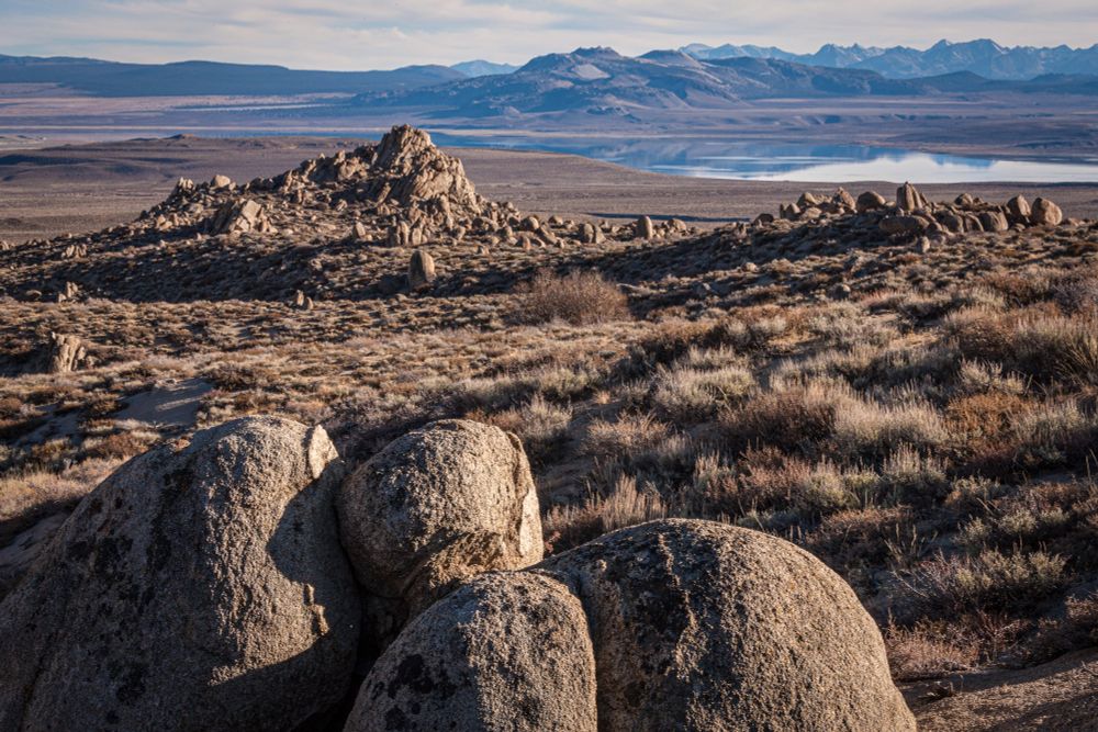 Daytime color photograph of a landscape, four warm brown smooth granite rocks in the foreground, brown grasses and rocks behind including a peak of rocks in the mid-distance.  Beyond that a smooth reflective lake near the top of the image, reflecting mountains and the rounded hills of the Mono-Inyo Craters, with a very pale blue, slightly clouded sky.