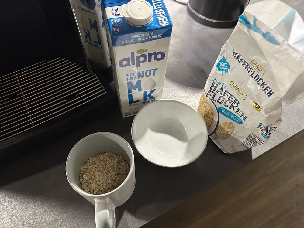 Eine Tasse und Schüssel stehen neben einer Packung Hafermilch und einer Tüte Haferflocken. Es wurden Haferflocken in die Tasse gefüllt statt in die Schüssel. 