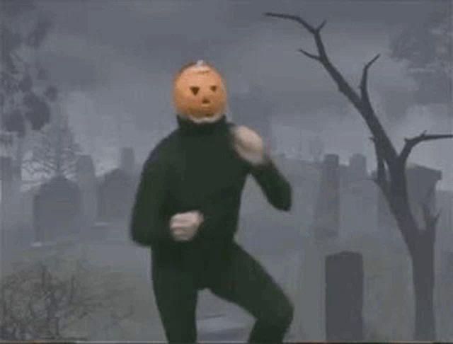 a man in a black suit with a pumpkin on his head is standing in a cemetery .