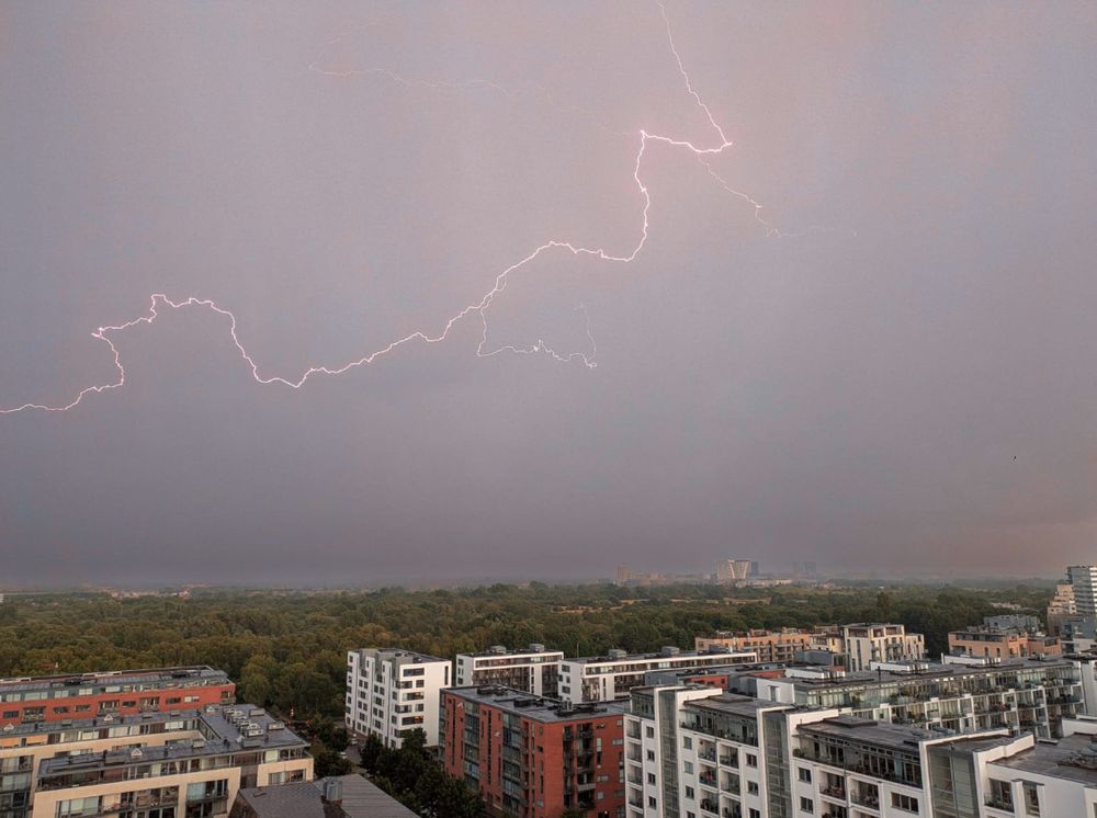 Lighting races across the sky above a series of buildings and, in the farther distance, trees