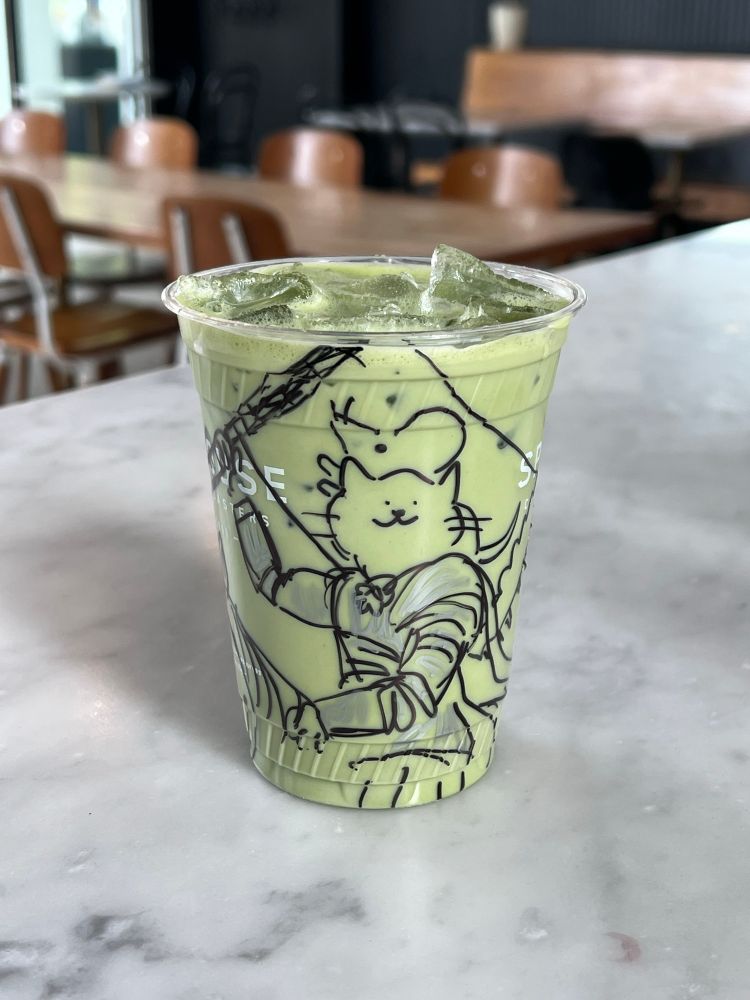 a photo of a to-go iced cup (matcha) with a sharpie drawing of a cat in armor riding a horse, holding a flag with a mouse on it, and a smile on it’s face. the cat’s armor is filled in with silver sharpie. 