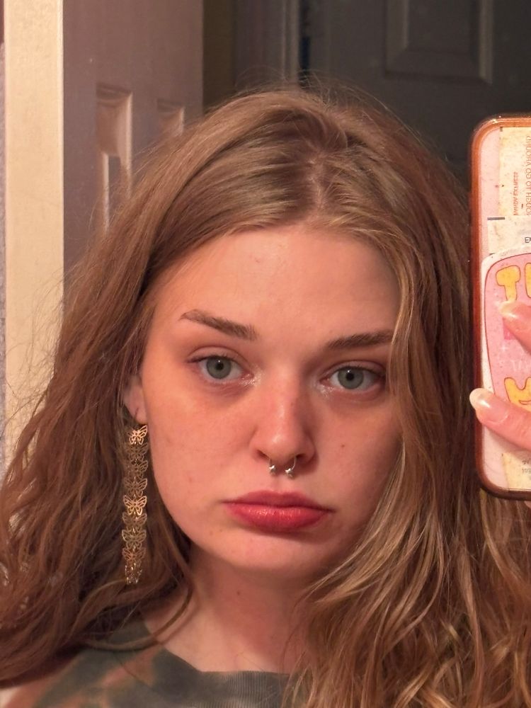a mirror selfie of merde, they are making eye contact with themselves not looking at the camera, they have wavy blonde hair and a butterfly chain earring, they have a neutral face 