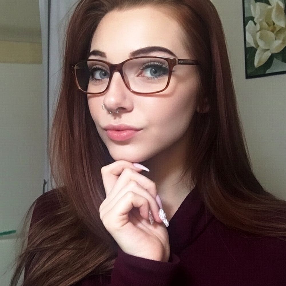 A young woman with reddish-brown hair and blue eyes is wearing brown glasses and a septum piercing, posing while resting her chin on her hand.