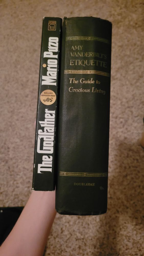 two books being held together, the godfather is on the left and is about half the size of a book with a black spine with gold lettering, titled 'Amy Vanderbilt's Ettiquette: The Guide to Gracious Living'
