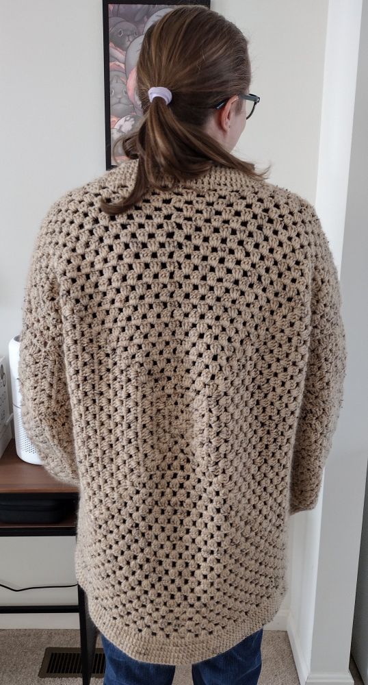 A person wearing a tan crochet cardigan standing to see the back of the cardigan.